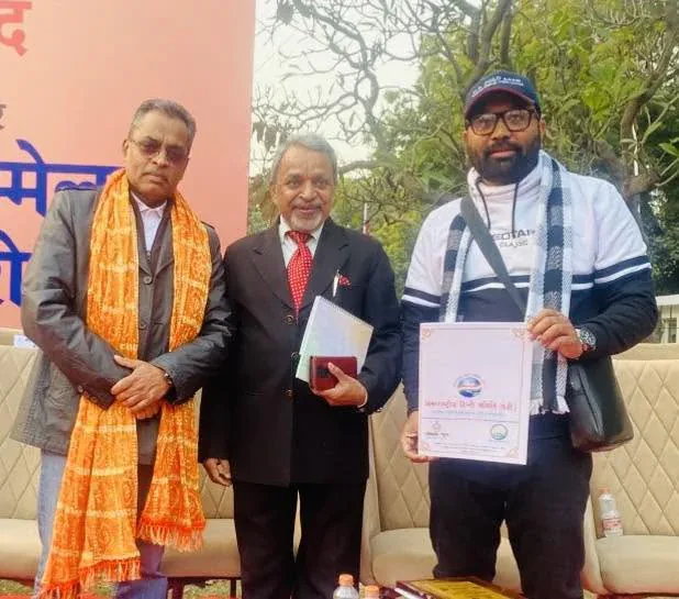 Vijay Kishor Tiwari holding a certificate, standing with two guests at a Hindi sammelan