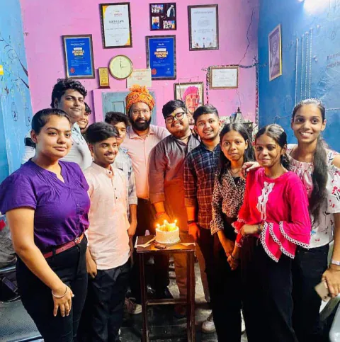 Vijay sir, in a saffron turban, cutting a cake with his students in front of the awards wall